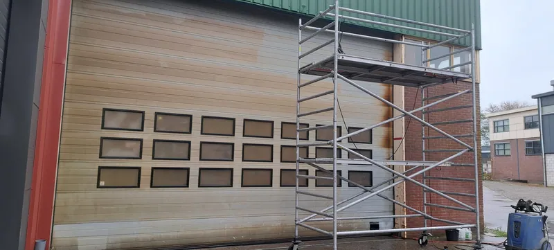 Bedrijfspand met een verweerde houten gevel en een lichte garagedeur, met een rolsteiger ervoor.