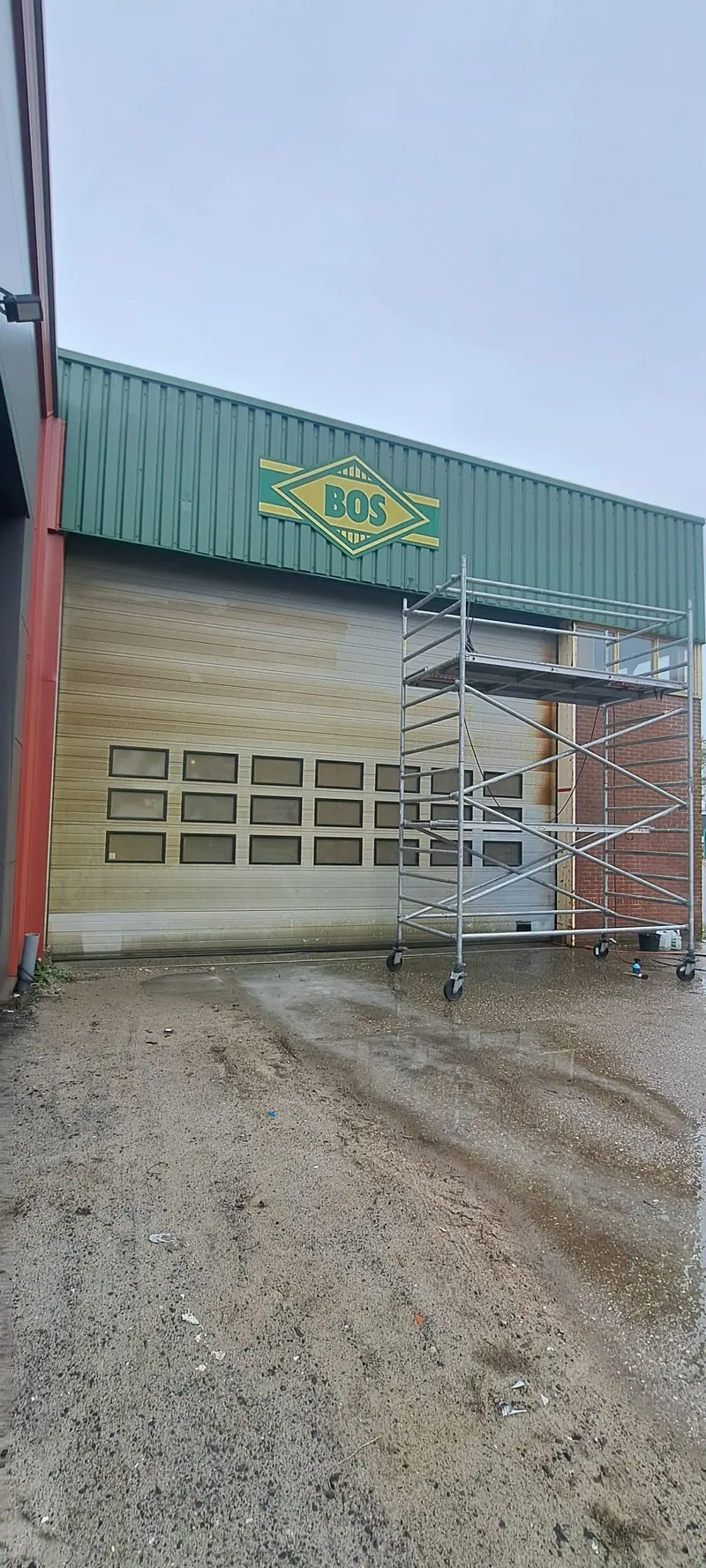 Bedrijfspand met een crèmekleurige garagedeur en een rolsteiger ervoor.
