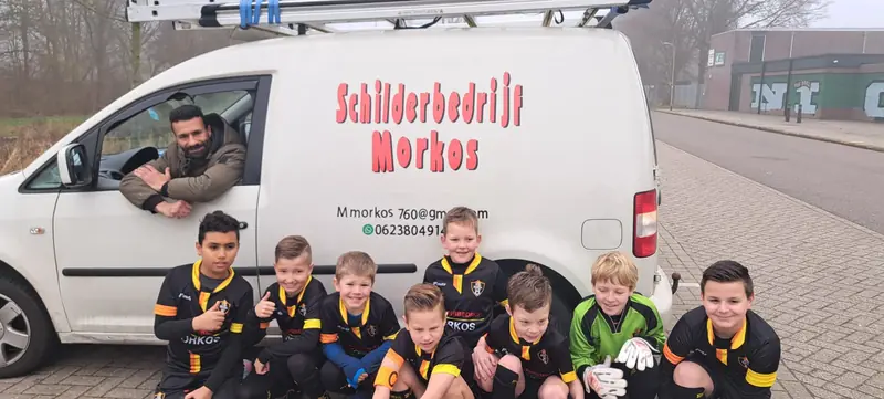 Milad Morkos leunt uit zijn bedrijfsbus, het jeugdelftal in tenue zit ervoor gehurkt.