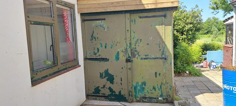 Groene garagedeuren met bladderende verf en een verweerd kozijn ernaast.