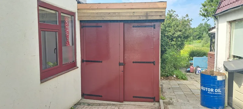Dezelfde dubbele garagedeuren en het raamkozijn ernaast, nu diep roodbruin geschilderd.