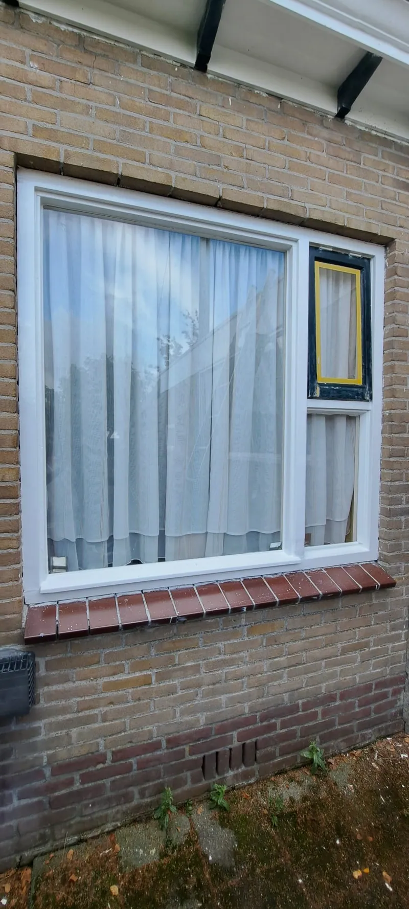 Wit kozijn met een geel geschilderd draairaampje in een lichte gevel.