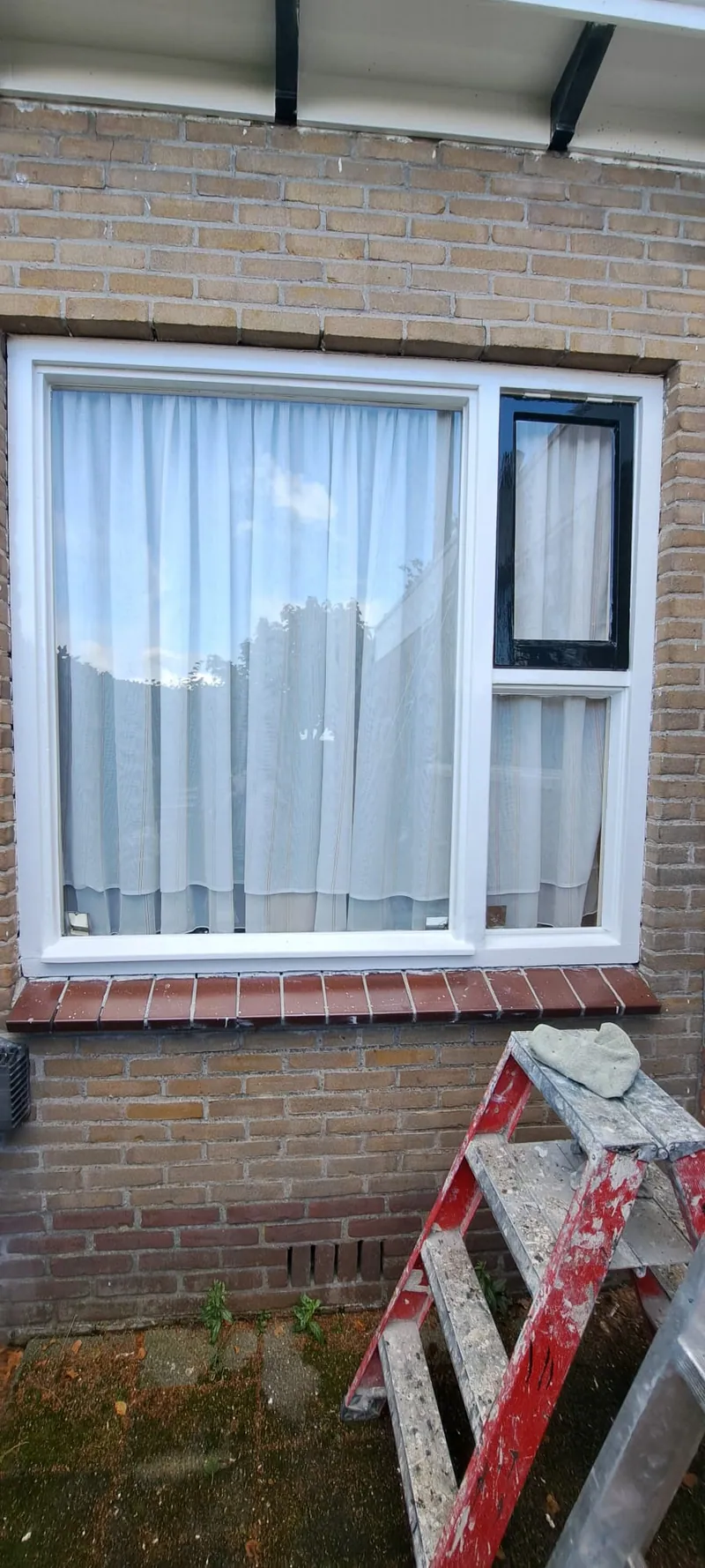 Wit kozijn met een zwart draairaam in een bakstenen gevel.