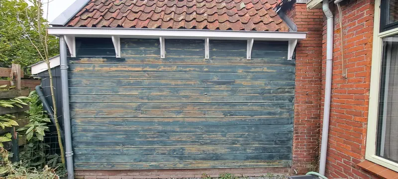 Houten aanbouw met afgebladderde blauwgroene verf naast een rode bakstenen muur.