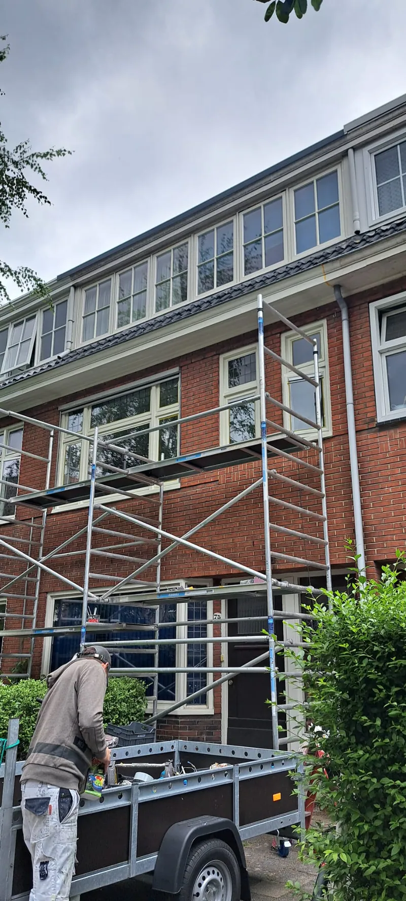 Steiger tegen een woning, met de aanhanger van het schildersbedrijf op de oprit.