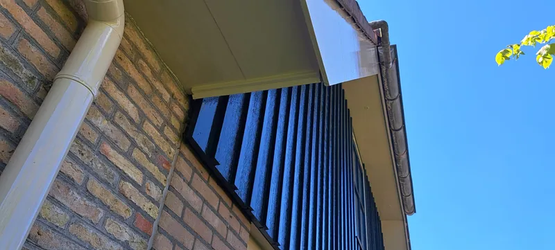 Blauw geschilderde houten topgevel met een witte goot tegen een bakstenen gevel.