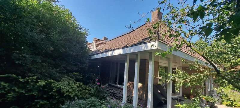 Woning met een veranda, half achter de bomen in de tuin.